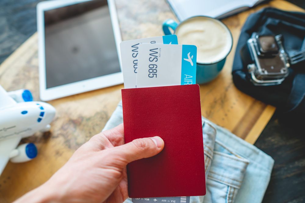 Ruka drži crveni pasoš sa avionskim kartama, dok se na stolu nalaze tablet, šoljica kafe, fotoaparat i model aviona